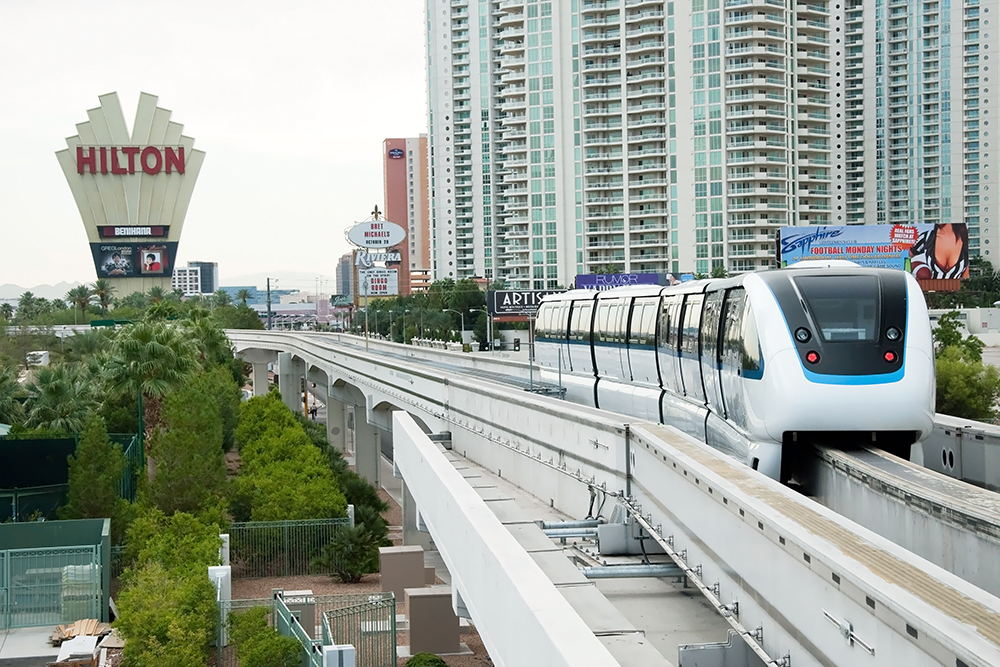A white monorail travels on an elevated track with high-rise buildings and trees in the background.